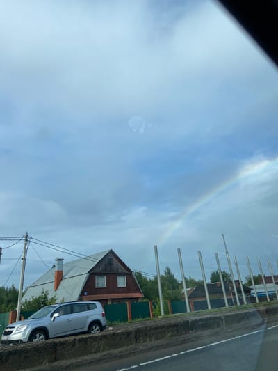 In Tarasovo, Russia, under a cloudy sky with a visible rainbow, a silver Chevrolet Orlando SUV is parked on the side of a road. The vehicle is positioned in front of a large, multi-story house with a distinctive asymmetric roofline and a prominent chimney. A green fence runs along the property line. The scene also includes utility poles with wires, and a row of metal poles in the background. The road surface in the foreground appears rough and worn.