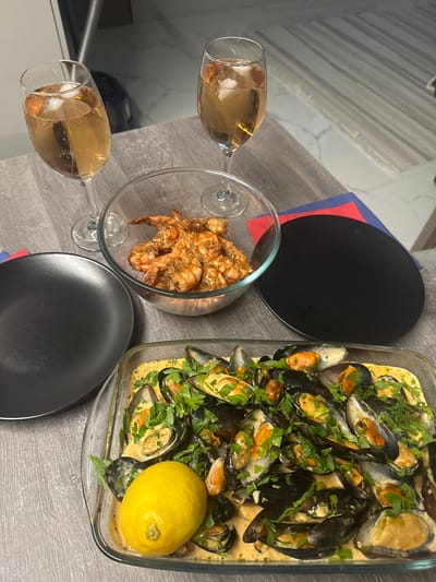 A close-up, eye-level, indoor shot depicts a table set for a meal, featuring seafood and drinks. The table has a grey wooden surface. In the foreground, a rectangular glass dish contains mussels in a creamy sauce, garnished with chopped parsley. A whole lemon sits in the lower-left corner of the dish. To the left of the dish is a matte black plate. Behind the plate, a glass bowl holds cooked shrimp. Two wine glasses filled with a light-colored liquid, possibly white wine or champagne, are visible in the background, each containing ice cubes. A black circular placemat is situated behind the glass bowl, resting on red and blue napkins. The background shows a blurred kitchen or dining area with tiled flooring and a metallic appliance. There are no people visible in the image. The scene appears to be a home setting, potentially in Sustaš, Montenegro, given the meal's presentation, suggesting a casual yet elegant dining experience.
