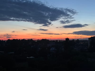 The image shows a twilight scene over a rural or suburban area, likely in the vicinity of Пратасевіцкі сельскі Савет, Belarus. The sky transitions from a deep blue at the top to vibrant orange and red hues near the horizon, indicating either sunrise or sunset. Dark clouds are scattered across the upper portion of the sky, contrasting with the lighter tones below.
In the foreground and midground, a dense silhouette of trees and buildings is visible. The structures appear to be low-rise residential buildings, with some rooftops and darker shapes suggesting houses and possibly some outbuildings. The overall impression is one of a quiet evening or early morning. There are no visible people or discernible actions taking place. The environment is characterized by its natural twilight lighting and the silhouetted landscape. The presence of what appear to be distant lights suggests some level of human habitation or infrastructure.