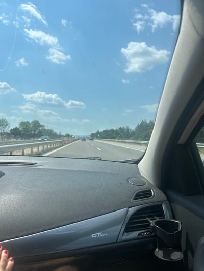 The image is taken from inside a car, looking out the windshield and to the right at the dashboard. The scene depicts a sunny day on a highway in Bulgaria. The sky is a clear blue with fluffy white clouds. Trees line the side of the road, and there is traffic visible ahead, with cars and what appears to be construction barriers. The dashboard is made of dark grey textured material, with a circular speaker vent and a car vent with horizontal slats. A black cup holder is attached to the car vent. There is some text visible on a silver trim piece on the dashboard, which reads "GT line". A person's hand with red painted fingernails is visible in the bottom right corner. The time of day appears to be daytime, and the weather is sunny and clear. The overall impression is of a typical road trip or commute.