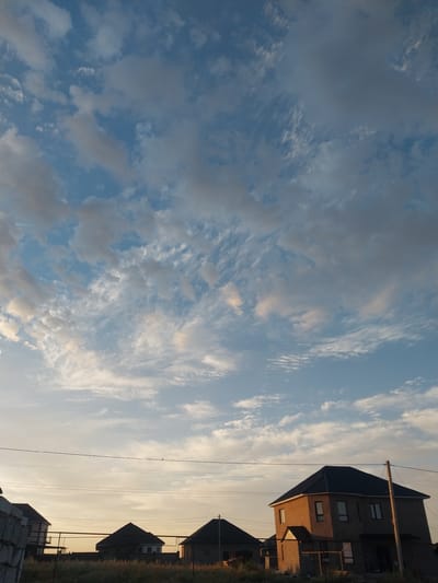 The image depicts a suburban or rural scene in Іргелі ауылдық округі, Kazakhstan, characterized by a dramatic sky with scattered clouds. Several houses, in various stages of construction or completion, are visible in the foreground and midground. These houses feature dark, angled roofs and light-colored walls. Utility wires stretch across the frame, supported by poles. The landscape is generally flat with sparse vegetation. The lighting suggests either early morning or late afternoon, with the sun casting a warm glow on some of the structures. No people are present in the image.