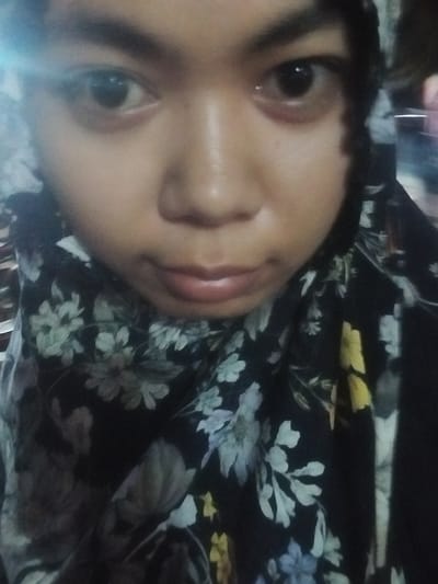 A close-up, low-angle, indoor, waist-up, medium shot features a young woman wearing a black headscarf with a floral pattern. The woman's face is visible, with her eyes looking directly at the camera. She has a light complexion and appears to be wearing light makeup. The background is blurred and dark, with hints of what might be a domestic setting. There are no observable interactions or actions. The image does not contain information related to the city of Tibojong, Indonesia.