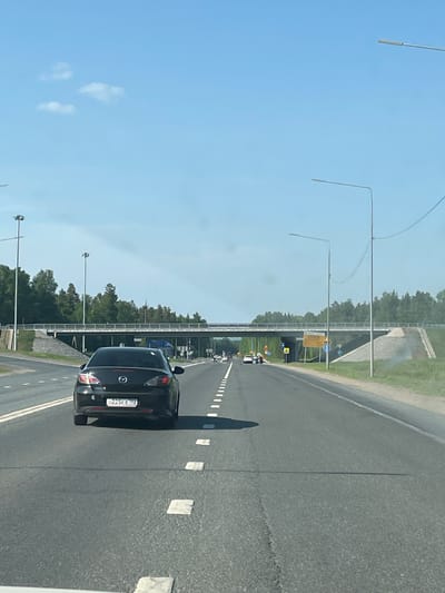 A photograph depicts a highway in what is presumed to be Severnyy, Russia, based on the vehicle license plate. A dark-colored Mazda is in the right lane of the highway. The scene is captured during daytime under a clear sky with sparse clouds. Streetlights line the side of the highway, which is surrounded by greenery. There are several vehicles in the distance. A bridge is visible over the highway. The license plate of the Mazda reads "c225ex 702".