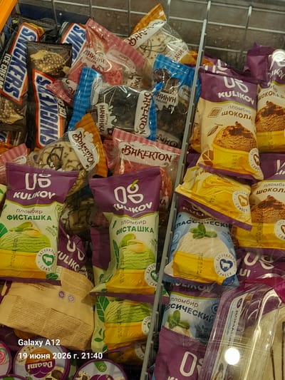 This is a close-up, overhead shot of a freezer display case filled with various ice cream products. The ice cream packages are predominantly in shades of purple, yellow, and green, with prominent branding and imagery of ice cream cones and flavors. Some identifiable brands include "KULIKOV" and "SNICKERS." The products are densely packed within the metal cage of the freezer. The lighting suggests an indoor commercial setting. The bottom left corner of the image shows a watermark indicating "Galaxy A12" and "19 июня 2026 г. 21:40", which are metadata from a mobile device and are not part of the scene itself. There are no people or interactions visible. The environment is clearly a commercial freezer, likely in a grocery store or supermarket. The city of Kara-Köl, Kyrgyzstan is not depicted or suggested by any visual elements in the image.