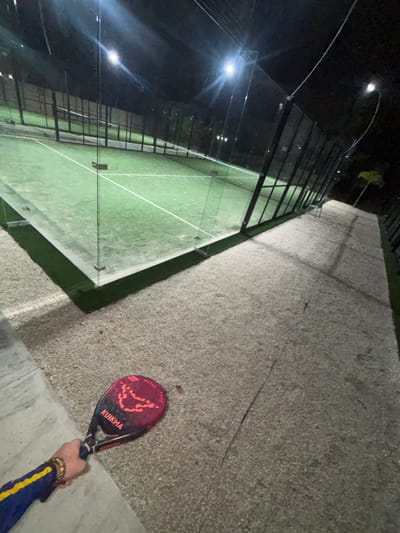 The image displays an outdoor padel tennis court at night, located in Privada Residencial Blue Cedar, Mexico. In the lower left foreground, the left arm and hand of an individual are visible, wearing a blue long-sleeved garment with a yellow stripe on the upper arm and a gold-colored bracelet on the wrist. The individual holds a KUIKMA brand padel racket, which is predominantly dark red with black accents and orange-red graphic designs, including the word "METAL". The racket is oriented downwards towards a light-colored gravel path that extends into the background along the right side of the frame. This path borders a narrow strip of green artificial turf that immediately surrounds the padel court. The court itself features a green artificial turf playing surface with distinct white boundary lines and is enclosed by clear glass panels forming its perimeter walls and dark mesh fencing above. The court is brightly illuminated by multiple artificial floodlights, visible as bright white points of light and reflections on the glass. A light-colored object, possibly a bench, is faintly visible beside the fence in the mid-ground on the right. In the distant background along the gravel path, a small, light-colored tree is present. The gravel path in the foreground exhibits several hairline cracks.