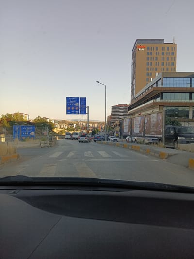 This media file captures a view from inside a car on a road in Yahşihan, Turkey. The scene depicts a moderately busy road with several vehicles, including cars and vans, moving in the same direction. On the right side of the road, there is a large, modern building with the prominent signage "podium kule" illuminated in red. Adjacent to this building are several large billboards with Turkish text and images, some of which are partially visible.

Further down the road, there are traffic signs, including those indicating speed limits and distance to various locations, with Turkish text such as "Ortalama Hiz Tespiti 4526 m" (Average Speed Detection 4526 m) and "EDS". The weather appears clear with a pale sky, suggesting it is either early morning or late afternoon. The overall setting is urban or semi-urban, with buildings and trees visible on either side of the road. The focus is on the road, traffic, and the surrounding infrastructure. There are no discernible people in the foreground or clearly visible in the vehicles. The mood is neutral, reflecting a common urban driving experience.