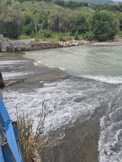 This is a medium-angle, eye-level, vertical shot of a river flowing over a small dam or weir. The water is turbulent and foamy in the foreground, indicating a rapid flow. The river is bordered by a concrete structure on the left, which appears to be part of the dam, and a rocky embankment with some gabion retaining walls on the right. Beyond the river, there is a dense forest covering a hillside, with some utility poles and wires visible among the trees. The sky is overcast, contributing to the muted colors of the scene. The image was taken in the city of Tekeli, Kazakhstan, likely at a recreational area or a point along the river where infrastructure has been built. There are no people or animals visible in the frame. The overall atmosphere is natural and serene, despite the presence of the man-made structures.