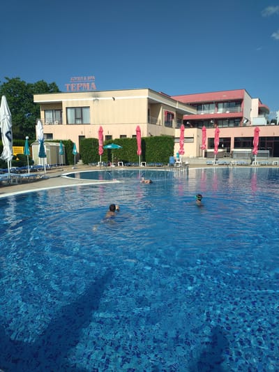 A bright, sunny day at a hotel and spa complex with a large blue swimming pool is depicted. The hotel building, painted in beige and pink, is visible in the background. A sign on the hotel reads "ХОТЕЛ & SPA ТЕРМА" which translates to "HOTEL & SPA TERMA". Several pink and green umbrellas are arranged around the pool area, along with lounge chairs. Three individuals are in the swimming pool: one adult male is swimming, and two children, one wearing goggles, are also in the water. The pool water is clear and blue, with reflections of the sky and surrounding objects visible on its surface. The environment appears to be a recreational area within a hotel complex, likely in Yagoda, Bulgaria, given the context.