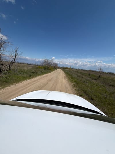 The image shows a dirt road stretching into the distance under a blue sky with scattered clouds. The road is flanked by grassy fields with bare trees. The hood of a white vehicle is visible in the foreground, suggesting the photo was taken from inside the car. The location is likely near Verkh. Lipovka, Russia, based on the prompt.