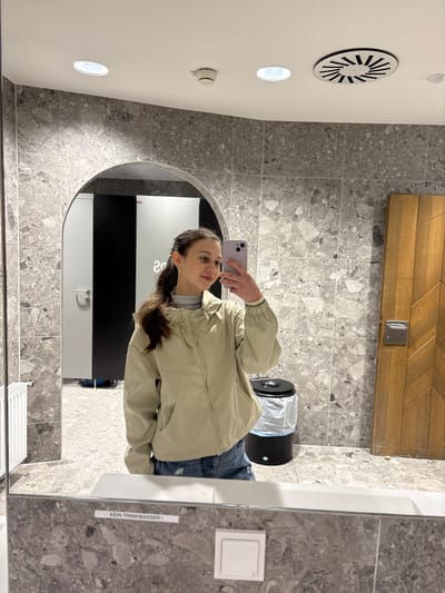 A young woman with dark brown hair tied in a ponytail is taking a mirror selfie in a modern restroom. She is wearing a light beige hooded jacket and denim shorts. She is holding a light purple smartphone in her right hand.

The restroom features grey marble-patterned tiles on the walls and floor. To the left of the woman, an arched doorway reveals a glimpse of stalls with black and white doors and a white radiator. A black trash can with a plastic liner is visible to her right. A wooden door with a chevron pattern is also on the right side of the frame. The ceiling has a circular vent and a recessed light fixture. In the reflection of the mirror, a white button and a label that reads "KEIN TRINKWASSER !" are visible. The overall impression is a clean and contemporary public restroom. The location in the city of Ehrwald, Austria is not discernible from the image.