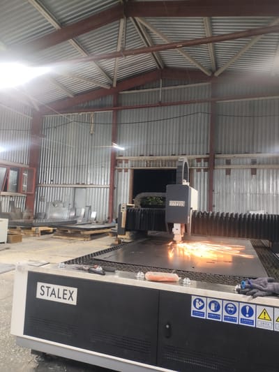 In a workshop in Dangir, Uzbekistan, a large industrial laser cutting machine is actively engaged in cutting a metal sheet. Sparks are flying from the point of contact, illuminating the immediate area. The machine has "STALEX" branding on its front panel. Several sheets of metal are stacked on pallets in the background, suggesting ongoing fabrication work. The workshop has a corrugated metal roof supported by steel beams and corrugated metal siding. Various tools, including pliers and a pair of scissors, are resting on the machine's surface, along with a pair of gloves and a small orange object, possibly a marker or a stylus. The overall environment appears to be a functional industrial space.