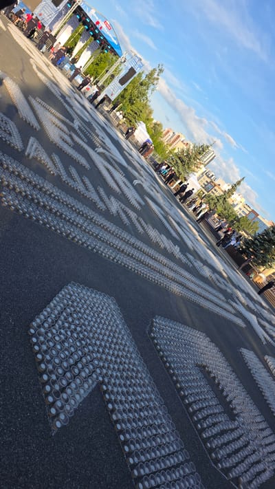 The image shows an outdoor event in the city of Дачное, Russia, likely a public celebration or festival. The foreground is dominated by a large-scale art installation or display made of many small, clear plastic cups arranged on the ground to form large numbers. The cups appear to be arranged in a grid pattern to create the shape of digits. In the background, a stage is set up with a large banner, and there are trees and buildings visible, suggesting an urban environment. Several people are present, some standing near the stage and others walking around the area. The sky is blue with some clouds, and the lighting suggests it is daytime, likely late afternoon due to the angle of the sun casting long shadows. The overall scene depicts a festive outdoor gathering.
