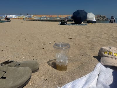 The image captures a beach scene in Toseong-myeon, South Korea, featuring personal items in the foreground and people in the background.

In the foreground, on the sandy beach, are a pair of grey sandals, a clear plastic cup containing ice and liquid, and a tan-colored baseball cap resting on a white towel. The sand is coarse and light brown.

In the midground, several people are scattered across the beach. A person sits in a chair near a low wall. Farther back, there are two people sitting under sun umbrellas, one dark blue and one white. A person stands near the water's edge.

The background consists of a light blue sky. Between the beach and the sky, there is a low wall with painted murals. Beyond the wall is a breakwater constructed of large concrete blocks.