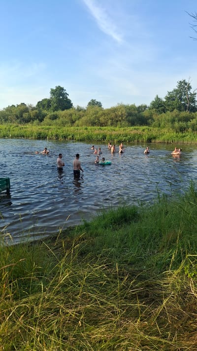 A group of approximately 15 people are swimming and wading in a wide, calm river. The water is dark and reflects the clear blue sky, which has a faint white streak of clouds. The riverbanks are lined with dense, green vegetation and trees. Two men stand in the water in the foreground, facing away from the camera. To their right, a child is visible in a green inflatable ring. Further out in the river, other individuals are scattered, some swimming, others standing or sitting in the water. A green object, possibly a bench or part of a structure, is partially visible on the left edge of the frame. The scene appears to be a recreational area in a natural setting, likely on a sunny day.