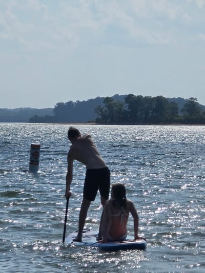 Two people are on a paddleboard in a body of water. A man is standing, holding a paddle. A young girl is sitting on the paddleboard. The water is choppy and reflects the sunlight. In the background, there is a tree line and a hazy sky. A buoy is visible in the water to the left of the paddleboard. This scene appears to be taking place on Lake Lanier, a reservoir in north Georgia, which is located in or near Flowery Branch, United States of America.