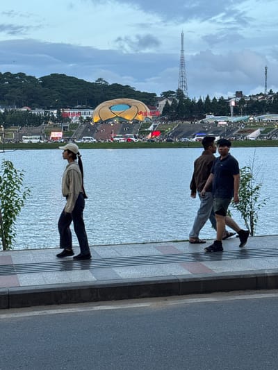 The image captures a waterfront promenade in Xuan Huong Lake, Da Lat, Vietnam. In the foreground, a young woman with a ponytail, wearing a beige cap and a tan jacket over dark pants, walks along the paved walkway. To her right, two men walk in the same direction. The man closer to the camera is wearing a dark blue polo shirt and camouflage shorts, with dark athletic shoes. The man behind him wears a brown jacket and light gray pants, with sandals. The walkway is bordered by a body of water on the left, reflecting the overcast sky. In the background, across the lake, is a vibrant cityscape featuring a large, lotus-shaped building with illuminated golden panels, stairs leading up to it, and dense green hills topped with trees. Two tall communication towers are visible on the horizon, one significantly taller than the other. The scene appears to be in the late afternoon or early evening, with a cloudy sky casting a soft light.