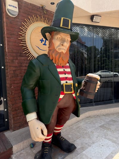 An outdoor statue of a leprechaun stands in front of a building with red brick walls. The leprechaun is wearing a green hat, a green jacket over a red and white striped shirt, brown pants, and black shoes with yellow laces. It is holding a wooden mug of frothy beer in its right hand. Behind the leprechaun, there is a large circular wooden decoration on the brick wall with sunbeam-like wooden spokes. The building has large glass windows with a decorative star pattern. A white car is visible reflected in the windows. The ground is paved with stone tiles. The location is the entrance to a business, likely a pub or restaurant, in Denizli, Turkey, suggested by the visible sign on the building that reads "Apax".