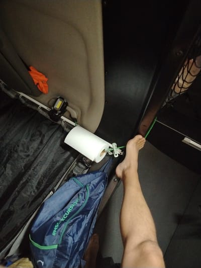 The image depicts the interior of a vehicle, possibly a truck or camper van, with a focus on the sleeping area. An individual's bare leg and foot are visible on the right side of the frame, positioned near a green cord that runs horizontally. This cord is attached to a roll of paper towels held by a clip, which is itself affixed to a white rod running along the wall. A dark blue bag with green trim, featuring the hashtag "#MSCXCURSION", is hanging below the paper towels. An orange object, possibly a piece of clothing, is draped over the top of the vehicle's interior lining on the left. A portable work light with a yellow beam is attached to the white rod, illuminating the area. The environment appears to be a confined space, suggesting travel or a temporary living arrangement. The image does not contain any specific geographical markers to indicate the location in Michendorf, Germany.