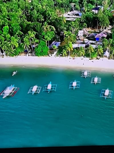 This is an aerial photograph of a tropical coastline. The left side of the image shows a dense green jungle with scattered buildings, indicative of a small town or resort. The buildings appear to be a mix of traditional and modern architecture, with some featuring colorful roofs. The jungle is lush and filled with palm trees.
The right side of the image depicts a calm, turquoise sea. Several small boats are moored in the water, close to a sandy beach that separates the jungle from the sea. The boats are of a traditional design, with outriggers, and are predominantly white with some blue and red accents. One boat has red detailing on its canopy. There are no people visible in or around the boats.
The scene suggests a sunny day, given the bright lighting and the clear water. The water is calm, implying fair weather conditions. The presence of the jungle and palm trees points to a warm, possibly humid climate. The overall impression is one of a tranquil coastal scene, likely a popular tourist destination. There is no visible text in the image.