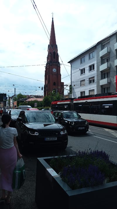 This is a daytime, outdoor shot captured in Nuremberg, Germany. The scene depicts a busy city street with a prominent red brick church tower topped with a cross, piercing the overcast sky. A modern tram with red and white detailing travels on the tracks to the right of the frame. Several black SUVs are stopped in traffic on the road. One SUV in the foreground displays a license plate reading "N RX 837", and another to its right has "N KB 1828". A woman, seen from the back, is walking on the sidewalk on the left, carrying a green and white shopping bag. She is wearing a white top and a long, light pink pleated skirt. A dark planter box filled with purple flowers is visible in the lower right quadrant of the image. The overall mood appears to be a normal, urban day with a sense of pedestrian and vehicle movement.