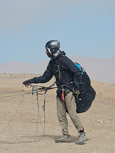 A man is paragliding in Alto Hospicio, Chile. He wears a dark jacket, khaki pants, hiking boots, a helmet, and goggles. The paragliding equipment is attached to him. The background shows a sandy landscape with some buildings in the distance and a blue sky above.