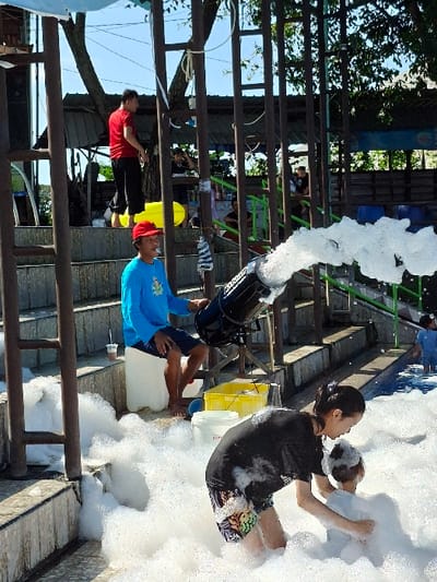 The scene is set in Kecamatan Tulung, Indonesia, at the coordinates -7.592 latitude and 110.65 longitude. A large amount of white foam is being blown by a machine in the center of the frame. It appears to be a foam party. A young adult woman in a black shirt and shorts is helping a young child play in the foam. There is a man standing to the left wearing a blue shirt and jeans. An empty white bucket is on the ground next to a yellow bucket. There are people in the background, but they are blurred. The location is a park-like setting with wooden structures in the background and a grassy area.