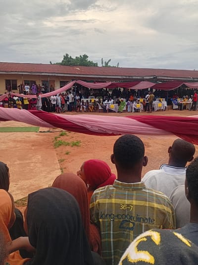 The image captures an outdoor event in Ogbogbo, Nigeria, under a heavily overcast sky. In the background, a single-story building with a weathered, reddish corrugated metal roof and light yellow walls features multiple windows and doorways. Several individuals are visible standing on a veranda in front of the building. Above the building to the left, green foliage is present.
The mid-ground is occupied by a large gathering. Red and light pink fabric draperies are suspended overhead, forming a decorative element or partial canopy. Beneath these drapes, numerous white tables and white plastic chairs are arranged. Many people are seated at these tables, some of which display small identification signs. A person in a light-colored garment stands near the tables, facing the seated attendees, suggesting they are addressing the group. Additional people are standing behind the seated crowd.
The immediate foreground shows the backs of several attendees observing the event. A person in the center wears a yellow and brown plaid shirt with "36-7" and "long-coordinate" printed on the back. To their left, an individual wears a black head covering, while another further left wears an orange head covering. To the right, a person in a light grey long-sleeved shirt with a white collar and blue stripe is visible, with white earphones. Other partially visible individuals wear red and reddish-brown head coverings. The ground consists of reddish-brown earth with sparse patches of green vegetation and a concrete section on the left.