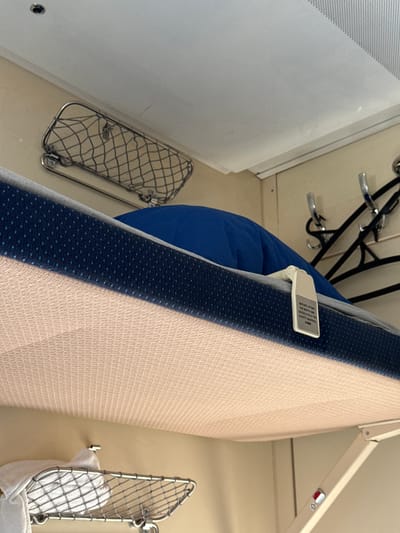 The image appears to be taken inside a train compartment. The angle is slightly skewed, looking upwards and towards the right. A bunk bed with a light peach-colored mattress and navy blue patterned trim is visible on the right side. The bunk appears to be in a folded position. A white tag is affixed to the side. Below the bunk is a small wire mesh basket with a white cloth or towel in it. To the left is a blue beanbag or cushion, with a coat rack behind it. Adjacent to the coat rack is a white wall with ventilation grids. Below the beanbag, a similar wire basket is mounted on the wall. The environment appears to be compact and utilitarian, typical of train interiors. There is no verifiable evidence of location within the city of Kirpichnoye, Russia.