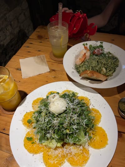 This image depicts a dining scene at a restaurant in 마포구, South Korea. The focal point is a rough-hewn wooden table laden with food and beverages under dim, ambient lighting.
Two main dishes are present:
1. A large white plate in the foreground features a salad composed of circular orange slices bordering a mound of mixed greens, topped with two balls of fresh mozzarella cheese, and generously garnished with grated cheese and black pepper.
2. A second white plate in the background contains green pesto pasta, garnished with two cooked shrimp, microgreens, a single half-cherry tomato, and a slice of toasted bread. This dish is also sprinkled with grated cheese and black pepper.
Beverages include a clear glass containing an orange-yellow iced drink on the left, and a tall clear glass with a lighter yellow iced drink and a clear plastic straw, resting on a cork coaster, positioned centrally.
Other objects on the table include a crumpled, damp white paper napkin situated between the two drink glasses. To the far right, a small metallic bowl contains green pickle slices. In the upper right background, a red, lobster-shaped handbag is partially visible, held by a person whose arm and hand are also partially visible.
The background consists of a dark, out-of-focus stone or brick wall, indicating an interior setting.