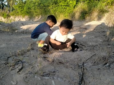 Two young children are playing in a sandy area in Meunasahkrueng, Indonesia. One child is squatting and wearing a blue long-sleeved shirt and light blue shorts with yellow and green shoes. The other child is sitting on the ground, wearing a white t-shirt and black pants with black shoes, holding a small stick. The ground is covered with sand, scattered dry leaves, and small dry grass patches. Behind them is a sandy embankment with green foliage on top. Coconut trees are visible in the background.