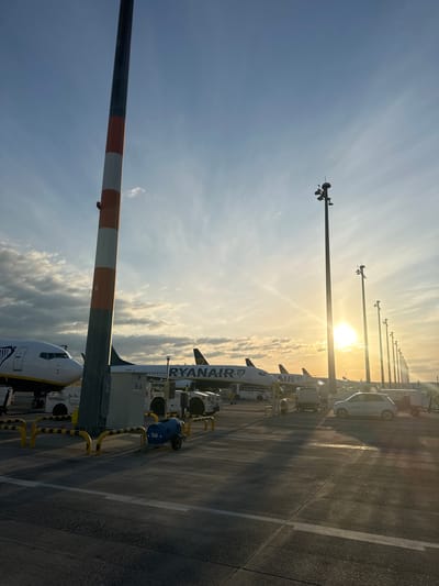 This image captures a sunset scene at an airport tarmac in Schönefeld, Germany. The sun is low on the horizon, casting long shadows across the asphalt. Several Ryanair airplanes are parked on the tarmac, their distinctive yellow and blue livery visible. In the foreground, a tall pole with orange and white stripes stands prominently. Various airport equipment, including service vehicles and a blue barrel on wheels, are also present. The sky is a mix of blue and grey clouds, with the sun creating a bright flare. A white car is parked on the right side of the image. The overall atmosphere is one of quiet activity at the end of the day.