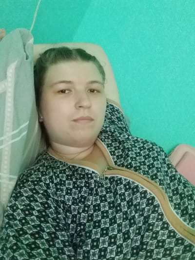 A woman lies on her side, facing the camera. She has her hair pulled back and is wearing a dark patterned robe with a tan zipper. A blue and white patterned blanket is behind her head. The wall behind her is painted a bright teal. The image is a selfie, taken indoors. No specific details about Kitskany, Moldova, are visible or suggested in the image.