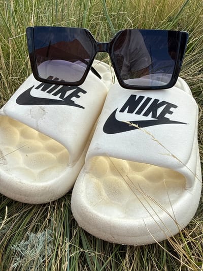 The image shows a pair of white Nike slide sandals with black Nike logos placed on dry, grassy terrain. A pair of black, oversized sunglasses rests on top of the sandals. The scene appears to be outdoors, likely in a natural setting with dry grass and some sparse greyish-green vegetation visible. The lighting suggests it is daytime, with the sun casting reflections on the sunglasses. There are no people visible in the image. The overall impression is one of relaxed, casual outdoor wear. The sandals and sunglasses are the primary objects of focus, set against a natural, somewhat arid backdrop. The prominent text visible is "NIKE" on both slides. The location context of Өндіріс, Kazakhstan, suggests a potentially dry, steppe-like environment, which aligns with the visual cues in the image.