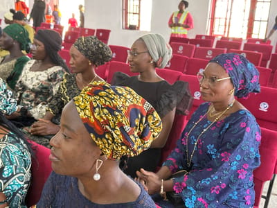 This image captures an indoor gathering in Goni Gara, Nigeria, featuring six adult women in the foreground and midground, with additional blurred figures in the background. Five women are seated in red upholstered chairs arranged in rows.
From left to right:
1. A woman, partially visible, wears a green and light-patterned garment.
2. A woman with dark skin and dark-framed glasses wears a patterned top featuring black, green, and white motifs, paired with a dark headwrap. She is looking towards the right of the frame.
3. A woman with dark skin wears a black top with ruffled short sleeves, a light grey headwrap, and clear-framed glasses. She has a subtle smile and a small stud earring on her visible ear. She is looking towards the right of the frame.
4. A woman with dark skin wears a dark blue top and skirt ensemble adorned with prominent three-dimensional floral embroidery in bright pink and light blue. She wears gold-rimmed glasses, a gold hoop earring, and multiple gold necklaces. Her head is covered with a dark blue patterned headwrap featuring similar pink and light blue floral designs. Her hands are clasped in front of her. She is looking towards the right of the frame.
5. A woman with dark skin, positioned in the lower right foreground, wears a dark blue lace top and a brightly patterned headwrap in yellow, orange, and dark blue. She wears a large dangle earring featuring a clear crystal triangular design suspending a white pearl. She is looking towards the right of the frame.
6. The sixth seated woman is partially obscured by the fifth woman's headwrap.
In the background, rows of empty red upholstered chairs are visible. A woman in the mid-background wears a bright pink top and a neon yellow safety vest, holding a dark object. Further back, blurred figures, some also appearing to wear orange safety vests, are discernible. The room features light-colored walls and tall, multi-paned windows with red frames on the far wall, allowing natural light into the space. The individuals appear to be attending an event or meeting, with most seated participants looking in the same general direction.