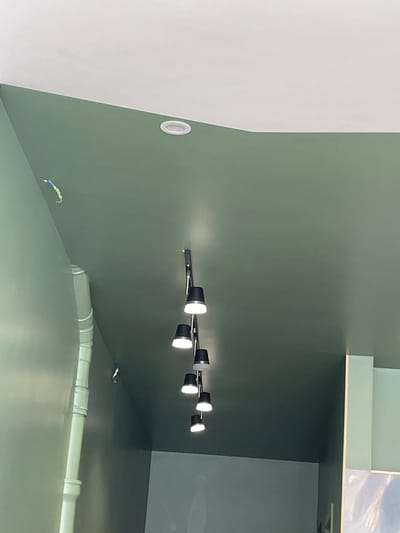 The image captures a perspective looking upwards towards a green ceiling and a portion of a white ceiling. A series of black pendant lights with white interiors hang from the green ceiling, illuminated. To the left, a white pipe runs vertically along a pale green wall. A small, brightly colored object, possibly a toy or decoration, is suspended from the ceiling. The scene appears to be an interior space, possibly a public building or a modernly designed room, given the architectural elements and lighting fixtures. The image does not contain any discernible elements that would allow for its specific location within Meaux, France.