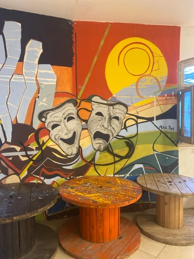 The image displays an indoor scene in Canning, Argentina, featuring a vibrant mural and three repurposed wooden cable spool tables. The large mural occupies the majority of the wall space, depicting an abstract composition. On the left side, light blue, vertically oriented shapes are set against a dark blue and orange background. The right side features a predominant red-orange background with a large yellow circle containing spiral motifs, and an abstract landscape of geometric shapes in green, blue, yellow, and brown. Centrally located on the mural are two grey theatrical masks, one depicting a laughing (comedy) expression and the other a crying (tragedy) expression, rendered with intricate details. A signature "MAIA PAN" with an associated year, possibly "2016," is visible on the lower right of the mural. Below the mural, from left to right, are three circular tables constructed from large wooden cable spools. The leftmost table is dark brown, the central table is painted orange with yellow paint splatters on its surface, and the rightmost table is a lighter, natural wood tone, partially visible. A window with multiple small blue glass panes is situated in the upper right corner of the scene, framed by a light-colored wall. No individuals are present in the image.