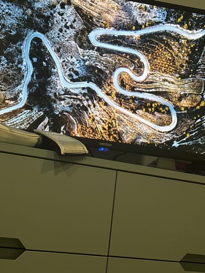 The image shows a portion of a monitor screen displaying an aerial view of a winding road through a mountainous terrain. The road is light-colored and contrasts with the dark and textured landscape. The landscape consists of varying shades of brown, black, and white, suggesting a mix of vegetation, rocks, and possibly snow. The monitor has a brand name "Weston" printed on its bezel. A partial view of a white and gray sandal is visible in front of the monitor. The background of the image consists of what appears to be a light-colored cabinet with vertical paneling. This does not appear to contain enough information to definitively state it is in Arinj, Armenia.