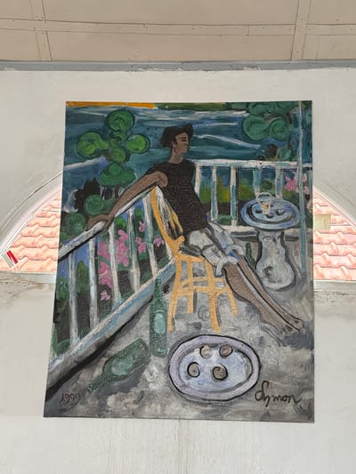 The painting, dated 1999 and signed "Symon," depicts a figure seated on a chair on a balcony. The figure, with dark hair and wearing a dark shirt and light shorts, leans on a railing. In front of the figure, a small table holds a drink. There are two green bottles and a plate with three circular objects on the ground in the foreground. The background suggests a landscape with water and trees. The painting is displayed on a white wall beneath a section of textured ceiling and adjacent to an arched window revealing red roof tiles and a small Indonesian flag. The setting appears to be a building in Tejakula, Indonesia.