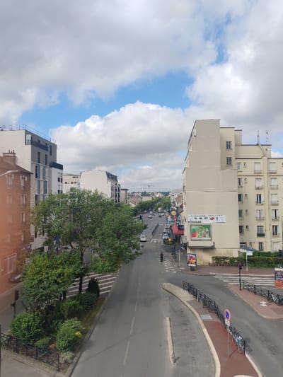 The scene depicts an urban street in Neuilly-Plaisance, France, viewed from an elevated perspective under a partly cloudy sky with patches of blue. A multi-lane, grey asphalt road occupies the central and lower portions of the image, leading into the distance.
In the foreground, the road forms an intersection with a curved, raised paved median on the right. Several bicycles are secured to a black metal fence on this median, and a red circular road sign with a white horizontal bar is mounted on a pole. On the left side of the road, a green, tree-lined median strip with bushes and a dark metal fence is visible. A multi-story building constructed of reddish-brown material, with ground-floor commercial establishments, is partially visible on the far left.
On the right side of the road, a tall, light beige multi-story building with numerous windows and balconies dominates. A large commercial banner displaying text "AUTO SALON" and "CHAMBRES D'HÔTES," along with images of vehicles and produce, is affixed to its facade. Below the banner, green shrubbery is planted. A red traffic light signal is present near the bottom right, and a partial yellow road sign is also visible.
In the mid-distance, a dark-colored scooter or motorcycle with a rider is observed traversing a zebra-striped pedestrian crossing. Multiple stationary vehicles, including at least two white cars, are visible along the street. Commercial storefronts are visible along the right side of the street in the mid-distance. The road recedes into the background, flanked by various multi-story buildings and trees. A construction crane is visible on the distant horizon.