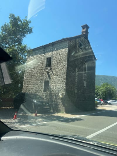 The image shows a stone building with two windows and a chimney on a clear day in Dražin Vrt, Montenegro. A black Nissan SUV is parked in front of the building, next to two orange traffic cones. The building is made of rough stone blocks. Trees are visible to the left and behind the building, with mountains in the distance. Several cars are parked in a lot to the right of the building. The image appears to be taken from inside a vehicle, as indicated by the windshield wipers and dashboard in the foreground.