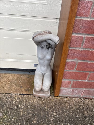 A supine stone sculpture of a human figure is positioned near a white door with a brown wooden lintel. A brick wall is above the lintel. The statue rests on a gray concrete surface. A black nameplate is affixed to the door below the statue. The location is Shrewsbury, United Kingdom.