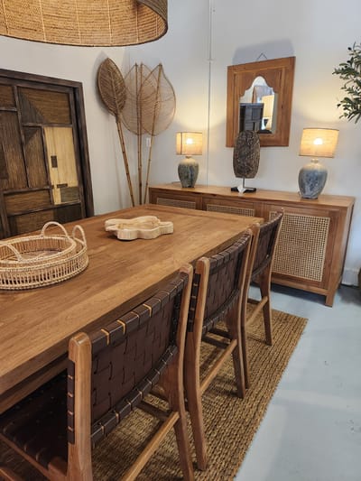 This media file showcases a well-lit room with a bohemian and naturalistic decor. The primary subjects are furniture items arranged in a setting that appears to be a retail display or a well-styled home.
In the foreground, a wooden dining table dominates, adorned with a small, carved wooden sculpture in the shape of a small animal, possibly a cat, and a woven basket. Two chairs with wooden frames and dark woven seats are positioned around the table, partially visible. A woven rug with a textured pattern is placed beneath the table and chairs.
Further back, a wooden cabinet with cane detailing stands against a white wall. Two lamps with square, textured shades and blue ceramic bases are placed on top of this cabinet, casting a warm glow. A framed mirror with a rustic wooden frame hangs on the wall above the cabinet, reflecting other elements of the room. Decorative items, including what appears to be a carved wooden mask or artifact with intricate patterns, are displayed on the wall.
To the left, a large, woven lamp shade or decorative piece hangs. To the right of the cabinet, a section of a wall and the floor are visible, suggesting a neutral color palette for the room. On the wall, near the mirror, decorative wall hangings made of woven materials and bamboo sticks, resembling large leaves or fans, are mounted. Below these, a section of a wooden door or panel with a geometric, inlaid design is visible.
The lighting suggests it could be daytime, with artificial light from the lamps also contributing to the overall illumination. There are no people visible, and the scene is static, implying a staged display. The overall impression is one of a calm, inviting, and texturally rich environment with a focus on natural materials. No visible text is present in the image. The location context is Maitland, United States of America.
