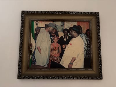 A framed photograph hangs on a white wall. The photograph depicts a group of people, including a young girl in traditional Igbo attire. Two men wearing hats stand on either side of the girl, appearing to be engaged in conversation. Other people stand behind them. A poster that reads "Means of Communication" is behind the group. The photo appears to be taken in Rumuokwuta, Nigeria.