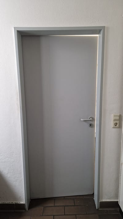 A plain, light grey interior door is centered in the frame, set within a grey doorframe. The door has a silver-colored door handle and a keyhole directly below it. To the right of the doorframe, a section of white wall is visible, featuring a light-colored electrical outlet with two sockets and a light switch. The floor beneath the door is tiled with dark brown rectangular tiles. The overall environment appears to be a neutral, indoor space. The lighting suggests an overhead light source is present, casting a bright illumination along the top gap between the door and the frame. No individuals are present in the image, and no specific actions are depicted. The scene is static and observational. The image was taken in Ichenhausen, Germany.