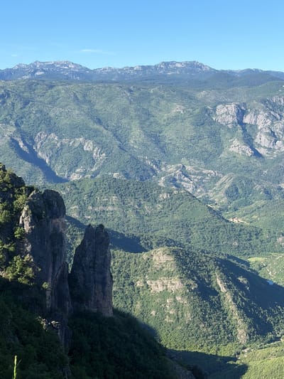 The image is a landscape photograph of a mountainous region in Kućišta, Montenegro. The foreground features two prominent, dark rock formations casting shadows on the lush green slopes. Beyond these formations, the landscape unfolds into a vast expanse of rolling green mountains covered in dense vegetation. The distant mountains are lighter in color, with some rocky outcrops visible, and they stretch towards a clear, bright blue sky. The scene depicts a natural, undeveloped environment, devoid of any people or man-made structures, except for a few scattered buildings visible in the valley far in the distance. The lighting suggests a sunny day, likely late morning or early afternoon, given the strong shadows.