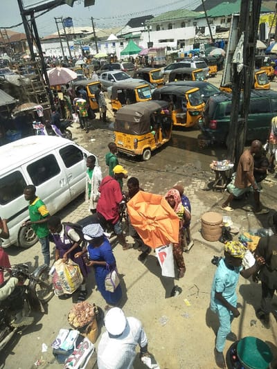 This image captures a congested outdoor market scene in Ajah, Nigeria, during daylight hours. The environment is characterized by a mix of pedestrian and vehicular traffic on a worn, unpaved or partially paved ground exhibiting visible wet patches. Overhead, a network of power lines and utility poles is present.
Approximately 25-30 individuals are visible throughout the frame, engaged in walking, standing, and vending. Their attire varies, including casual clothing, traditional garments, and what appears to be a blue school uniform worn by a person near the white van. A central figure is seen holding a large, open orange umbrella, possibly for shade or sale. Several individuals appear to be vendors, with goods displayed on small wooden stools or trays, such as packaged snacks, produce, and bottled drinks.
Vehicular traffic includes a prominent cluster of at least ten yellow and black auto-rickshaws (Keke Napep), some appearing stationary in a queue, others possibly navigating the street. A white van is parked or moving on the left, and a dark green SUV and a silver sedan are visible in the middle and background, contributing to the congestion. A motorcycle is partially visible on the left.
In the background, two to three-story buildings with light-colored facades and some green-colored roofs are present, along with market stalls featuring green and white awnings. A brown plastic barrel rests on the ground on the right side of the frame.