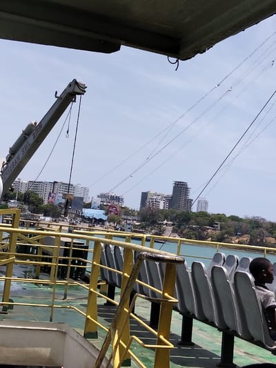The image depicts a daytime scene from the deck of a vessel, likely a ferry, with a view of the Mkomani ward skyline in Kenya.
In the immediate foreground, an overhead grey structure partially obstructs the top of the frame. Below this, a grey crane assembly, marked "SEMIZSAN SWL 2TKN," is positioned on the left side, extending upwards with a cable hanging vertically. A system of yellow metal railings forms the primary visual barrier across the mid-ground, indicating a passenger deck. To the right of the railings, a row of grey, individual, fixed-back seats is arranged, facing towards the right side of the frame. A young, dark-skinned male child is seated alone on the rightmost visible seat, wearing a striped shirt and facing right, with only his profile visible. The deck floor is a muted green color.
Beyond the vessel, a body of turquoise-blue water separates the ferry from the distant shoreline. Numerous thin, dark cables are strung across the clear blue sky, extending from the left to the right of the frame, some with small white insulators or markers.
The background reveals the urban landscape of Mkomani ward. Multiple multi-story buildings, varying in height and design, line the horizon, interspersed with patches of green vegetation and trees. Some structures appear to be residential or commercial complexes, and one prominent, darker multi-story building is visible towards the center-right. Closer to the water's edge on the land, indistinct structures, possibly smaller buildings, vehicles, and activity, can be perceived, though details are blurred due to distance. The sky is bright and clear with no discernible clouds, indicating fair weather conditions.