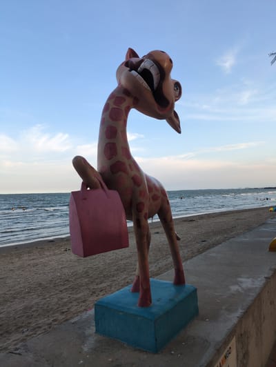 This image depicts a humorous statue of a giraffe at the beach in Hoằng Tiến Commune, Vietnam. The giraffe statue is pink with reddish-brown spots, has a wide, open mouth with exaggerated teeth, and appears to be holding a pink handbag in its right foreleg. It stands on a blue rectangular base on a concrete walkway that borders a sandy beach. The sea is visible in the background with gentle waves and a few people swimming. The sky is a pale blue with some wispy clouds. The overall scene suggests a tourist attraction or a whimsical decorative element in a public space.