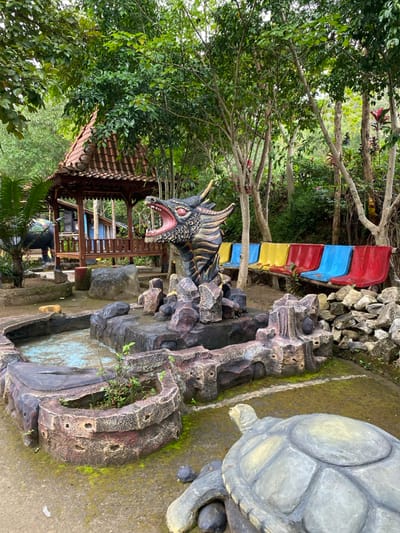 The image shows a small, artificial pond surrounded by rock structures. In the center of the pond, there is a sculpture of a dragon with its mouth open and its head tilted to the right. There are several rocks surrounding the pond, creating a sense of an enclosed space. A large turtle sculpture is located to the right of the pond. To the right of the pond, there is a row of colorful benches, made from wood and plastic. In the background, there is a wooden gazebo with a tiled roof. Lush green foliage surrounds the entire scene, creating a sense of natural beauty. The image is likely taken in an outdoor park, and it appears to be a popular tourist attraction. The exact location is a park in Kecamatan Manyaran, Indonesia, with the coordinates -7.826, 110.816.