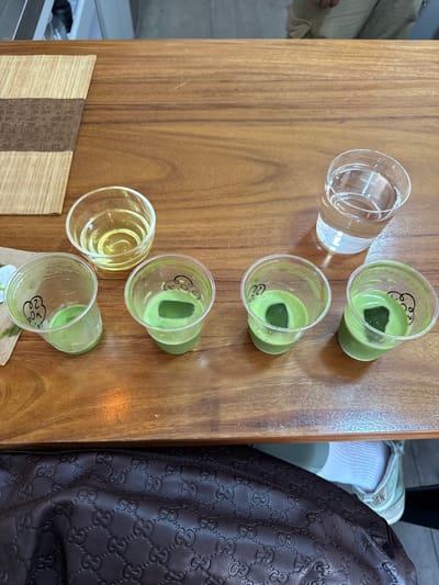 The image captures an overhead view of various drinks arranged on a polished wooden table, likely within a cafe or tea house in Guadalajara, Mexico. The scene suggests a relaxed gathering or tasting event.

The primary subjects are several beverages. Five small, clear plastic cups contain a vibrant green liquid, indicative of matcha, each featuring a single dark green ice cube and some foam on top. Two larger, clear glass tumblers are also present: one holds clear water, and another contains a yellowish liquid, possibly another type of tea or beverage.

In the bottom left corner, a small, light-colored bamboo mat or scroll adorned with intricate East Asian characters is visible, adding a cultural element to the setting.

On the right side of the frame, parts of a person's attire and accessories are visible. A dark brown, embossed leather bag, featuring a repeating "GG" logo pattern characteristic of Gucci, rests beside the table. Above it, a person's leg and foot are partially seen, wearing a white ribbed sock and a light-colored sneaker with the letters "DEN" visible on its side, possibly part of a brand name like "ADIDAS" (partially cropped). Another person's leg, clad in light-colored pants, is subtly visible on the far left edge of the image.

The lighting appears bright and even, typical of an indoor daytime setting. No specific time of day or weather conditions are discernible from the interior context. The overall impression is one of a casual, contemporary beverage experience.
